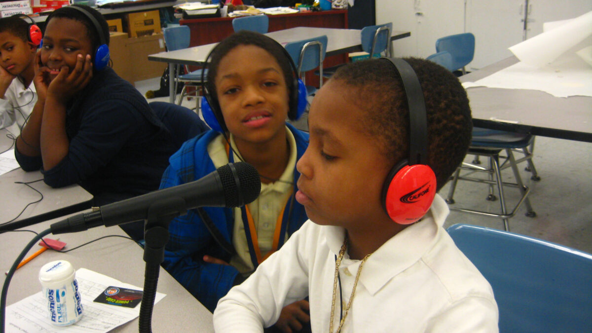 Four youth wearing headphones sitting at a white table, one speaking into a microphone