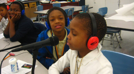 Four youth wearing headphones sitting at a white table, one speaking into a microphone