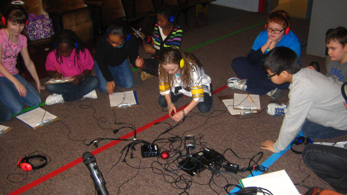 Eight youth, six wearing headphones, sitting on the floor around a microphone and a portable recorder
