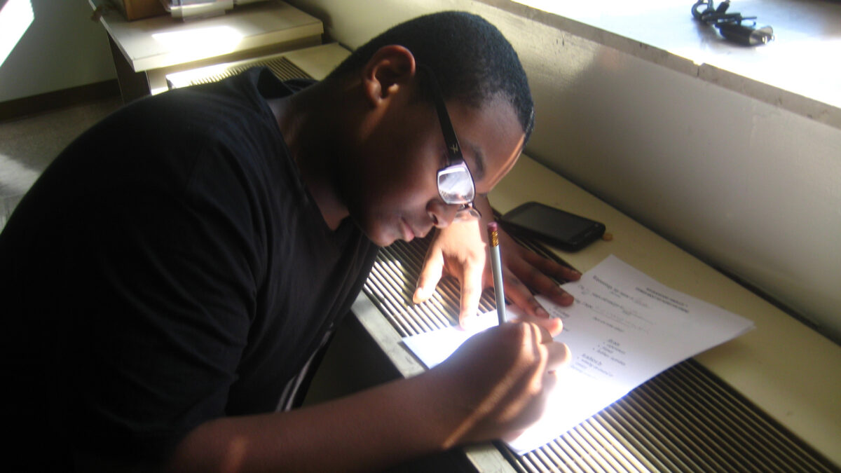 A youth sitting underneath a window writing on a piece of paper