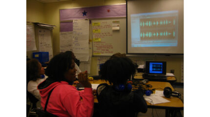 Three youth wearing headphones looking at audio editing software on a projected screen in a classroom.