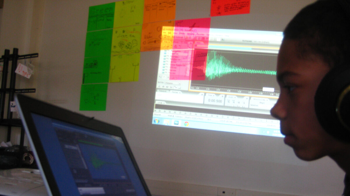 A youth wearing headphones looking at a laptop computer with a projected screen of audio editing software on a whiteboard in the background