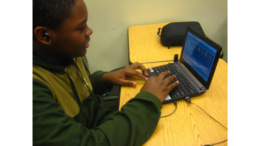 A youth wearing earbuds interacting with a laptop computer running audio editing software while sitting on a classroom desk