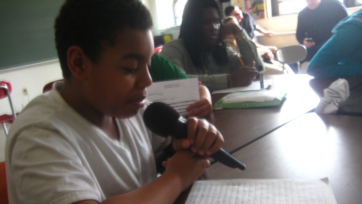 A youth holding and speaking into a microphone while looking at a piece of paper on a brown desk in a classroom