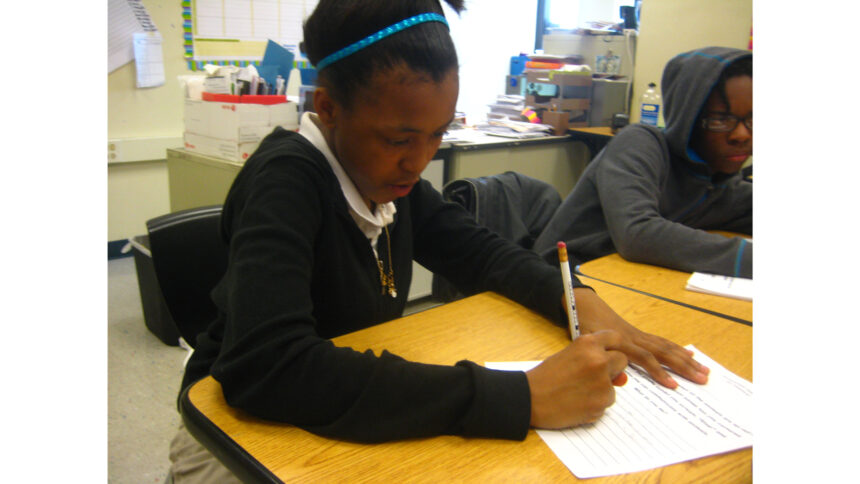 Two youth writing on pieces of paper sitting at wooden desks in a classroom