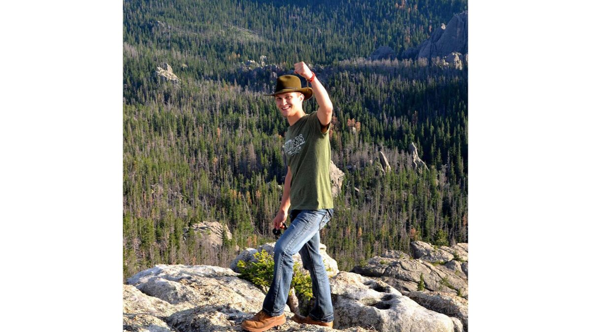 An adult holding up their fist while standing on rocks in front of a view of a forest