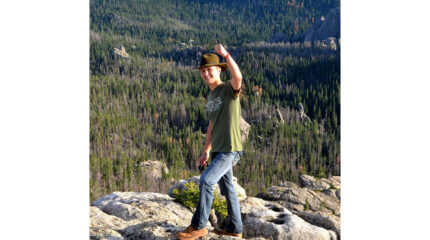 An adult holding up their fist while standing on rocks in front of a view of a forest