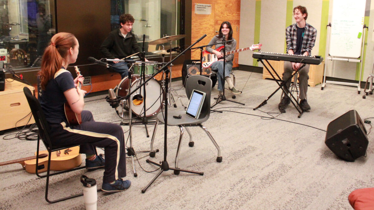 An adult sitting and holding a ukulele, an adult sitting at a drum set, an adult smiling and sitting while holding an electric bass, and an adult sitting at a piano keyboard