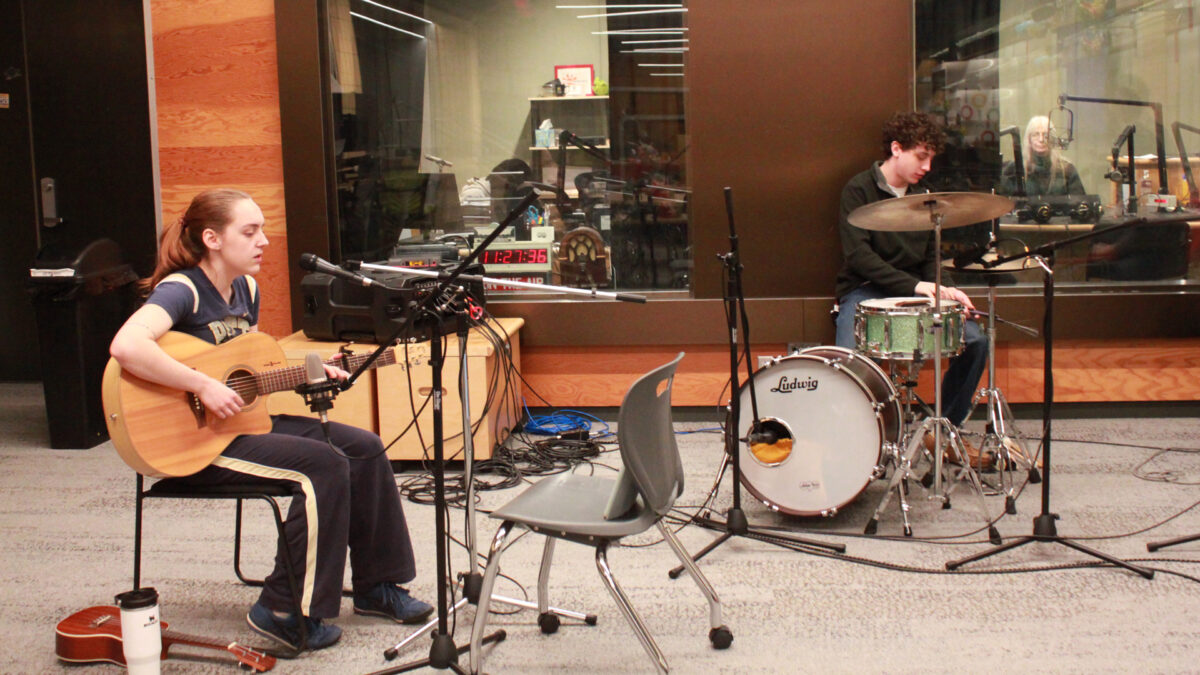 An adult sitting while playing an acoustic guitar and singing into a microphone and an adult playing a drum set