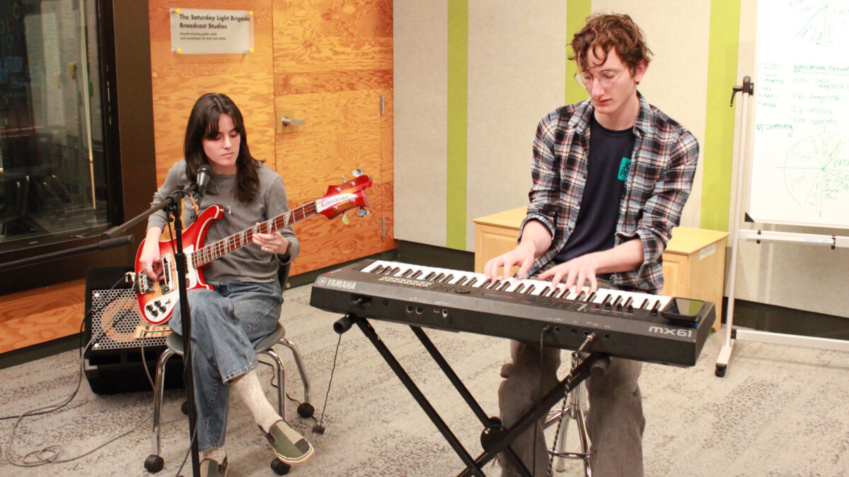 An adult sitting behind a microphone playing an electric bass and an adult playing a piano keyboard