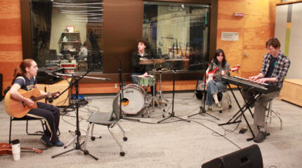 An adult sitting and singing into a microphone while playing an acoustic guitar, an adult playing a drum set, an adult sitting behind a microphone playing an electric bass, and an adult playing a piano keyboard