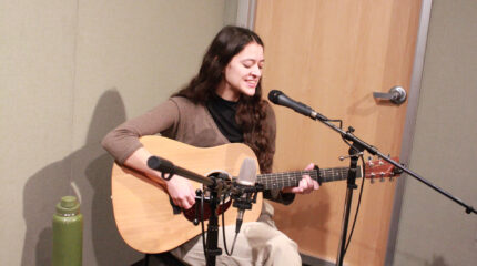 An adult singing into a microphone and playing an acoustic guitar on a radio studio