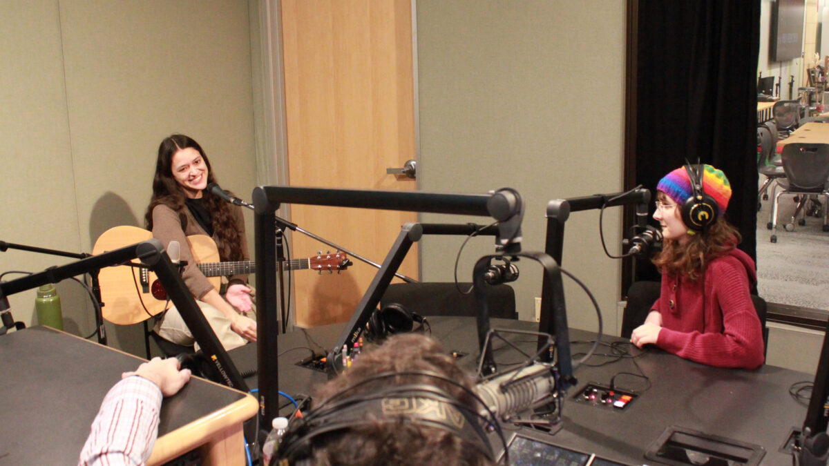 An adult smiling while sitting behind a microphone and holding an acoustic guitar and a teen wearing headphones sitting behind a microphone being interviewed by an adult wearing headphones sitting behind a microphone and a mixer in a radio studio