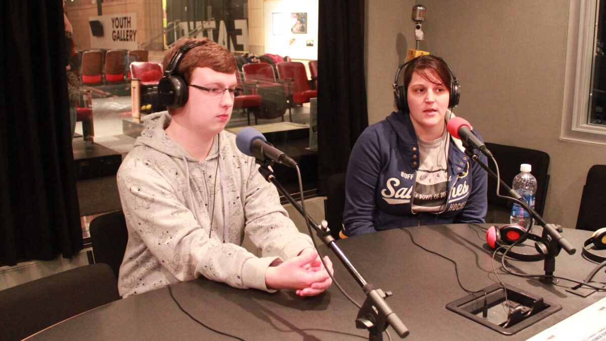 An adult and a teen wearing headphones speaking into microphones in a radio studio