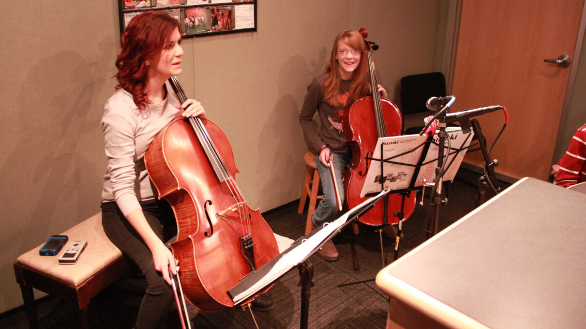 An adult and a youth holding cellos and sitting behind sheet music on music stands in a radio studio