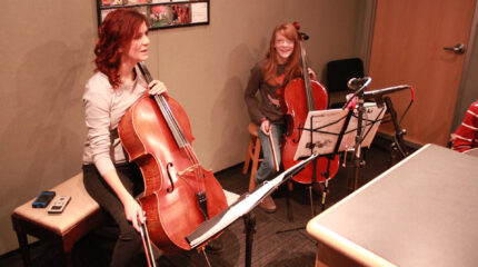 An adult and a youth holding cellos and sitting behind sheet music on music stands in a radio studio