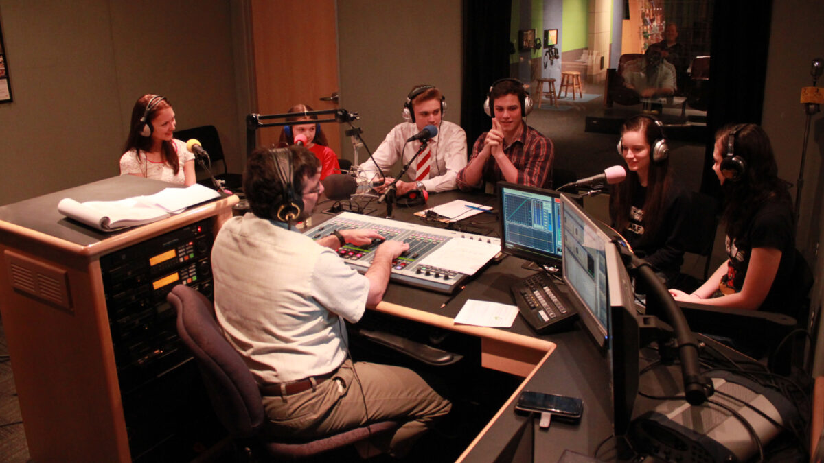 Six teens wearing headphones speaking into microphones being interviewed by an adult wearing headphones sitting behind a microphone and a mixer in a radio studio
