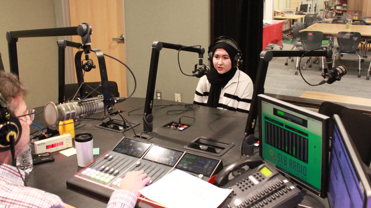 A teen wearing headphones speaking into a microphone being interviewed by an adult wearing headphones sitting behind a microphone and interacting with a mixer in a radio studio