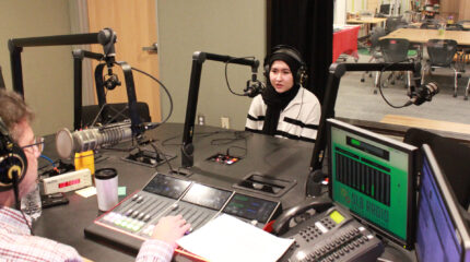 A teen wearing headphones speaking into a microphone being interviewed by an adult wearing headphones sitting behind a microphone and interacting with a mixer in a radio studio