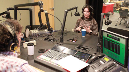 A young adult wearing headphones and speaking into a microphone being interviewed by an adult wearing headphones sitting behind a microphone and a mixer in a radio studio