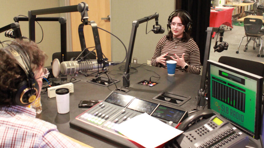 A young adult wearing headphones and speaking into a microphone being interviewed by an adult wearing headphones sitting behind a microphone and a mixer in a radio studio