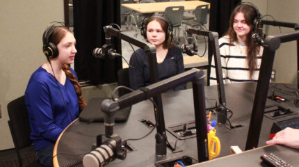 Three teens wearing headphones speaking into microphones being interviewed in a radio studio