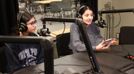 Two teens wearing headphones speaking into microphones in a radio studio