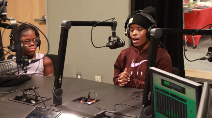 Two teens wearing headphones speaking into microphones in a radio studio