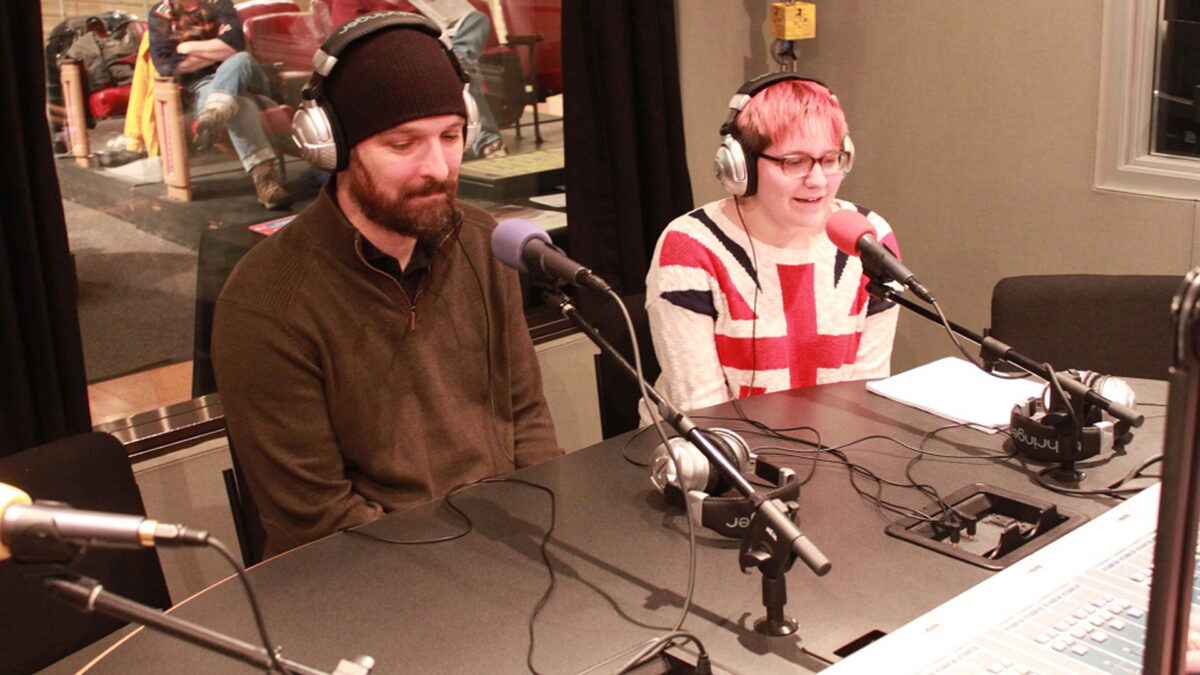 An adult and a teen wearing headphones speaking into microphones in a radio studio