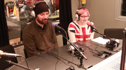 An adult and a teen wearing headphones speaking into microphones in a radio studio