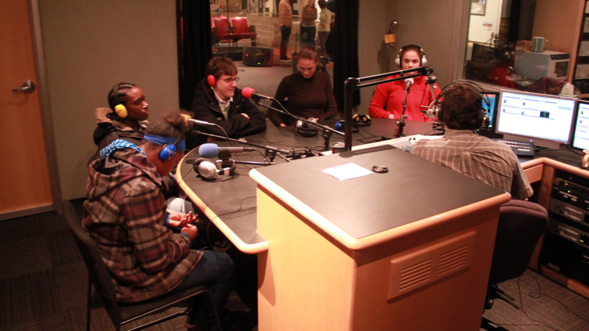 Five teens wearing headphones sitting behind microphones being interviewed by an adult wearing headphones sitting behind a microphone and a mixer in a radio studio