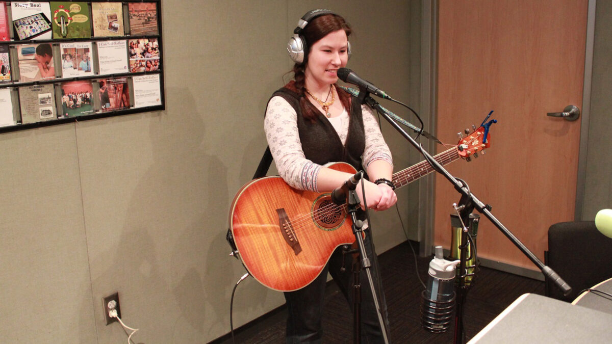 An adult wearing headphones holding an acoustic guitar and speaking into a microphone in a radio studio