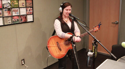 An adult wearing headphones holding an acoustic guitar and speaking into a microphone in a radio studio