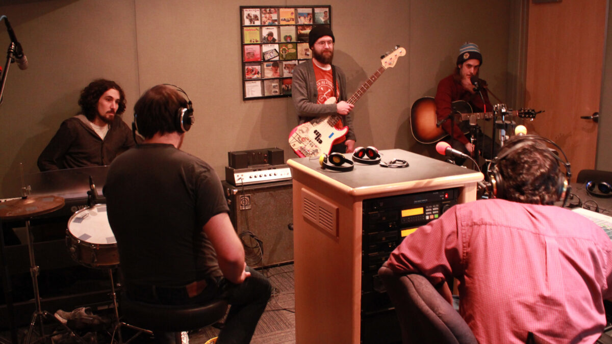 An adult sitting behind a piano keyboard, an adult sitting at a drum set, an adult holding an electric bass, and an adult sitting and holding a guitar and speaking into a microphone as an adult wearing headphones listens in a radio studio