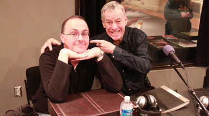 Two adults smiling while sitting behind microphones in a radio studio