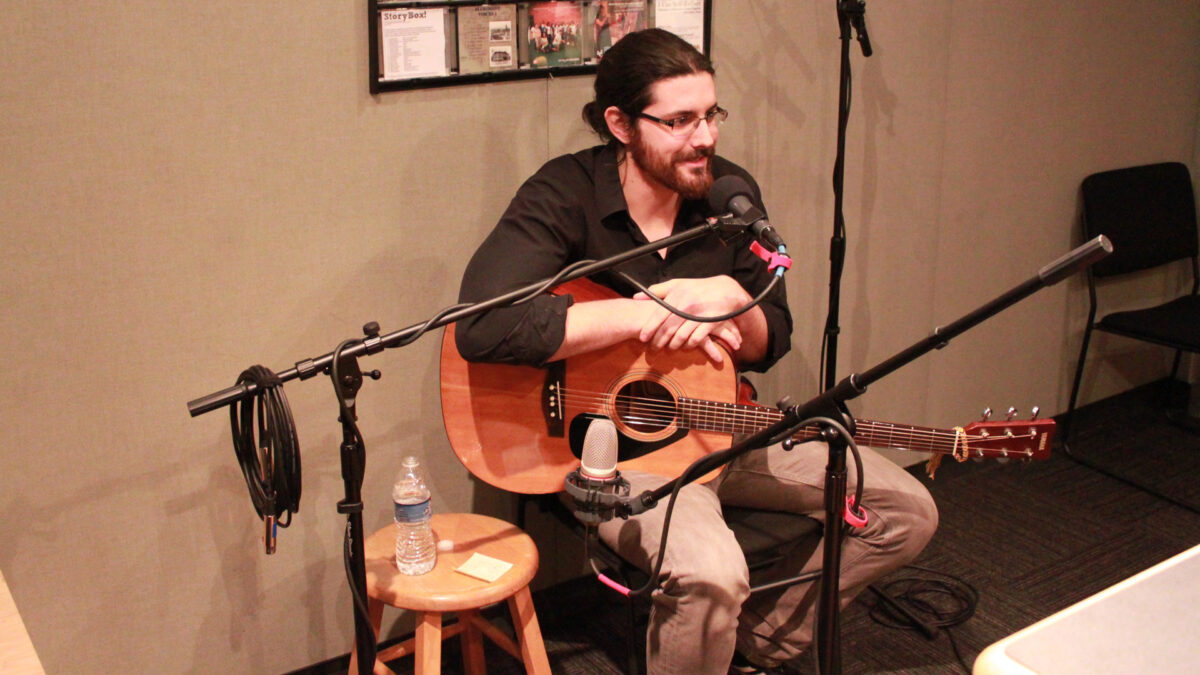 An adult sitting while holding an acoustic guitar and speaking into a microphone in a radio studio