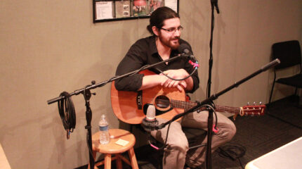 An adult sitting while holding an acoustic guitar and speaking into a microphone in a radio studio