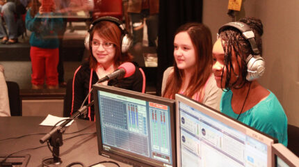 Three youth wearing headphones speaking into a microphone in a radio studio
