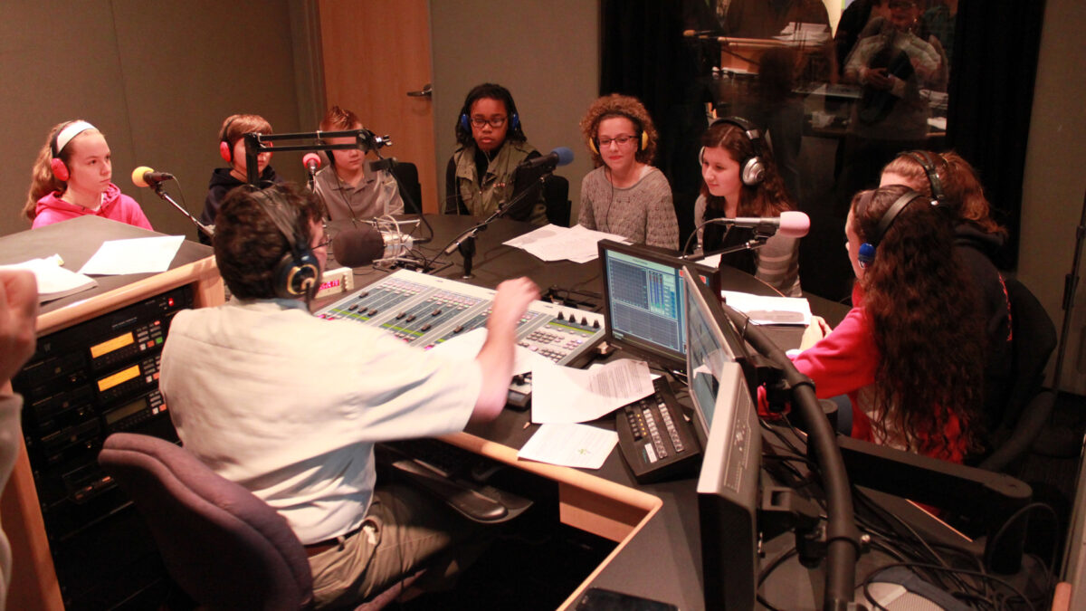 Eight youth wearing headphones and sitting behind microphones being interviewed by an adult wearing headphones sitting behind a microphone and a mixer in a radio studio