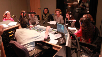 Eight youth wearing headphones and sitting behind microphones being interviewed by an adult wearing headphones sitting behind a microphone and a mixer in a radio studio