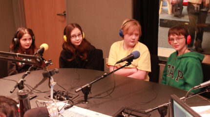 Four youth wearing headphones sitting behind microphones in a radio studio