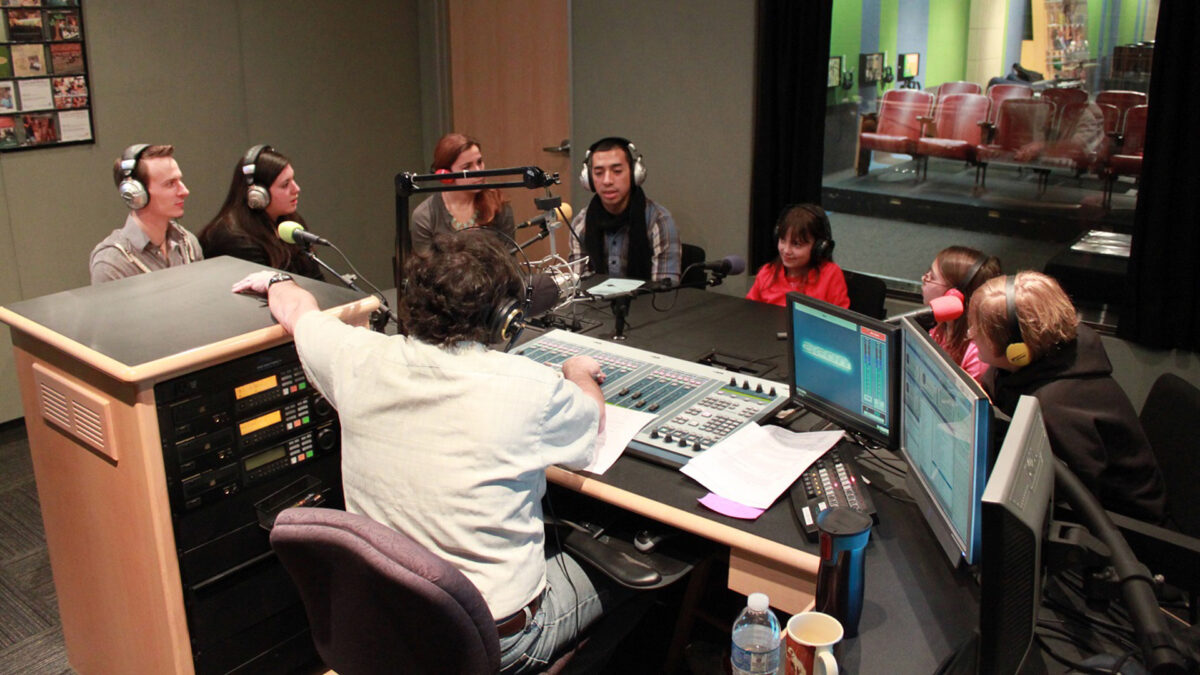 Four adults and three youth wearing headphones speaking into microphones being interviewed by an adult wearing headphones sitting behind a mixer and a microphone in a radio studio