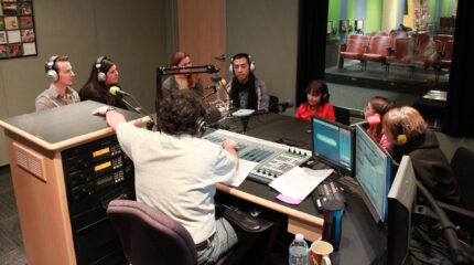 Four adults and three youth wearing headphones speaking into microphones being interviewed by an adult wearing headphones sitting behind a mixer and a microphone in a radio studio