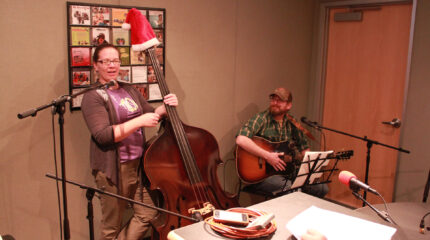 An adult holding an upright bass with a Santa hat on top and an adult sitting behind a microphone and sheet music on a music stand while holding an acoustic guitar in a radio studio