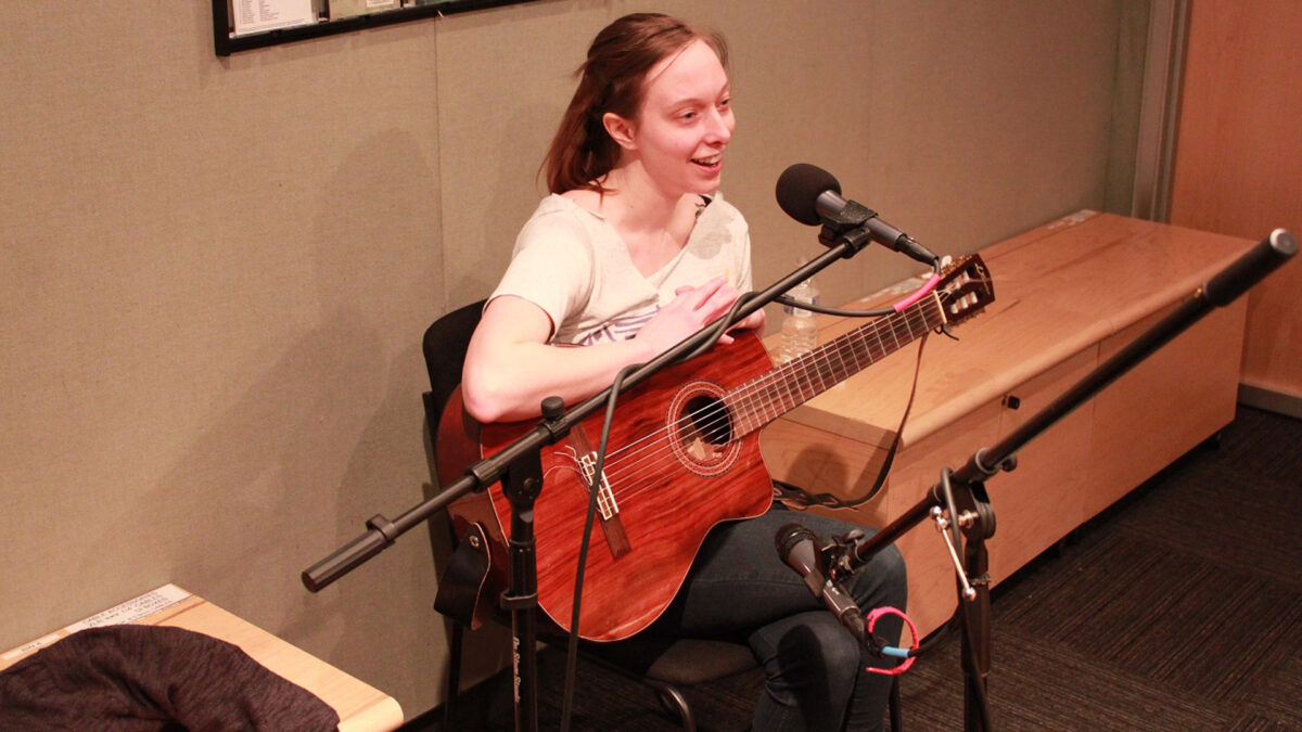 An adult sitting while holding an acoustic guitar and speaking into a microphone in a radio studio