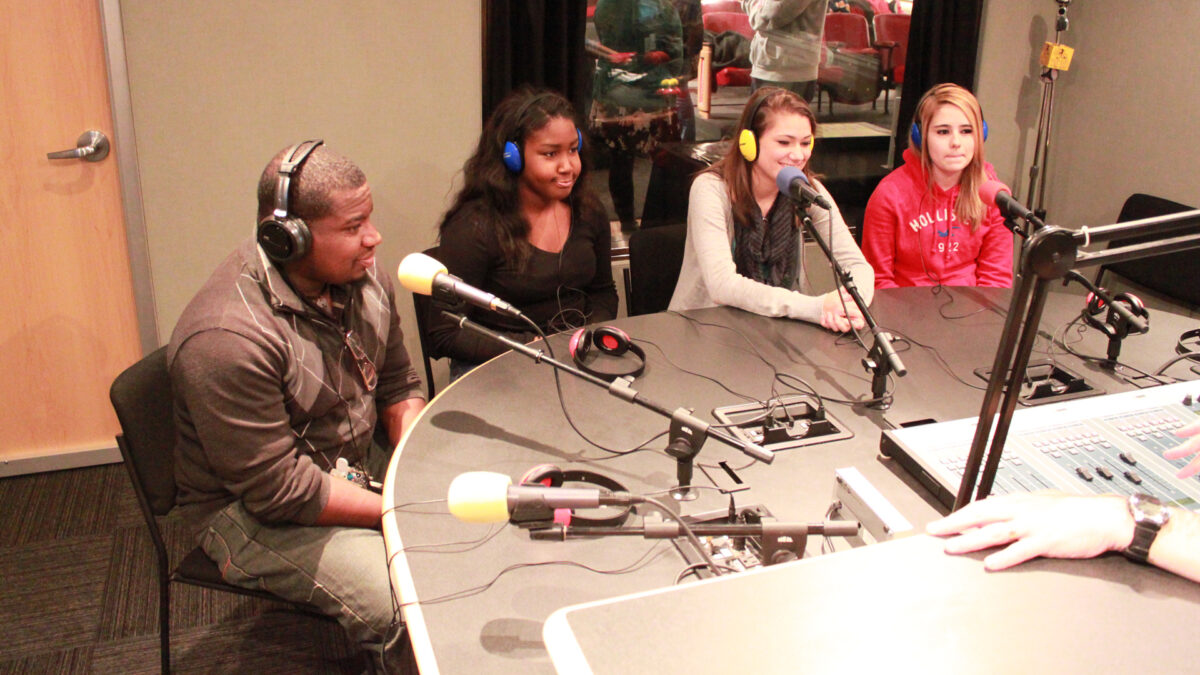 Four teens wearing headphones speaking into microphones in a radio studio