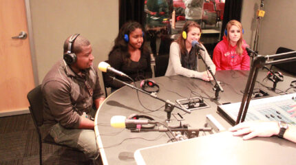 Four teens wearing headphones speaking into microphones in a radio studio