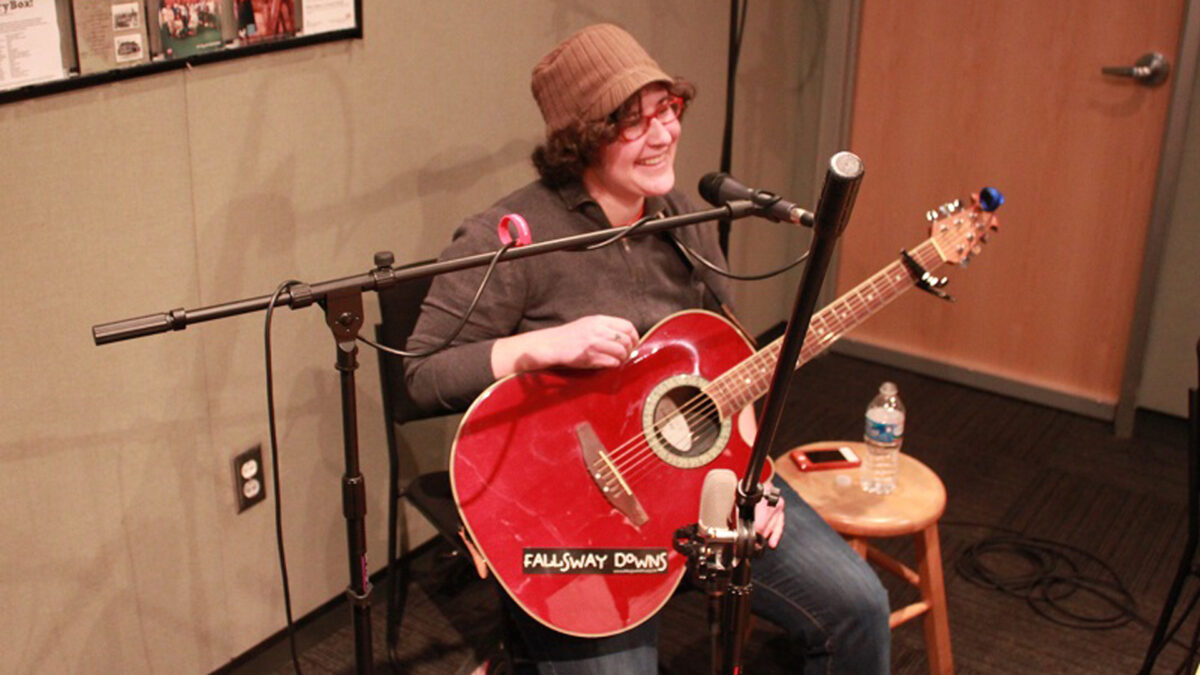 An adult sitting and holding an acoustic guitar while singing into a microphone in a radio studio