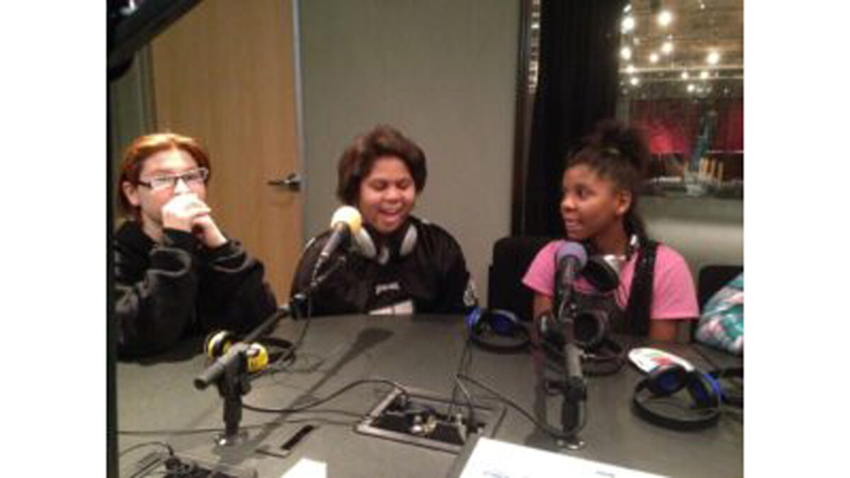 Three youth, two wearing headphones around their necks, speaking into microphones in a radio studio