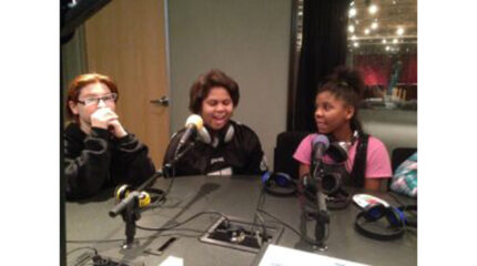 Three youth, two wearing headphones around their necks, speaking into microphones in a radio studio